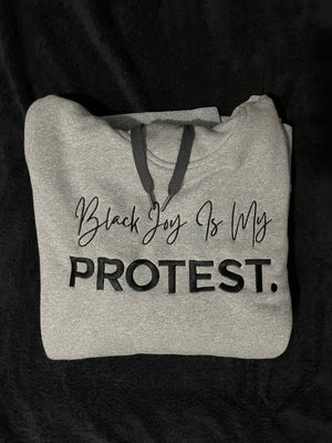 folded woman's fitted gray hoodie with black writing that says "black joy is my protest."