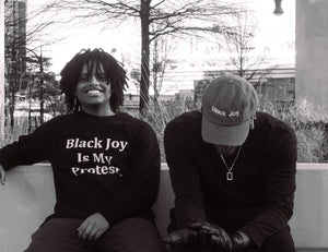 two Black men sitting on a bench. One with a "Black Joy Is My Protest Sweatshirt" and the other with a Black Joy hat on. Photo is in black and white
