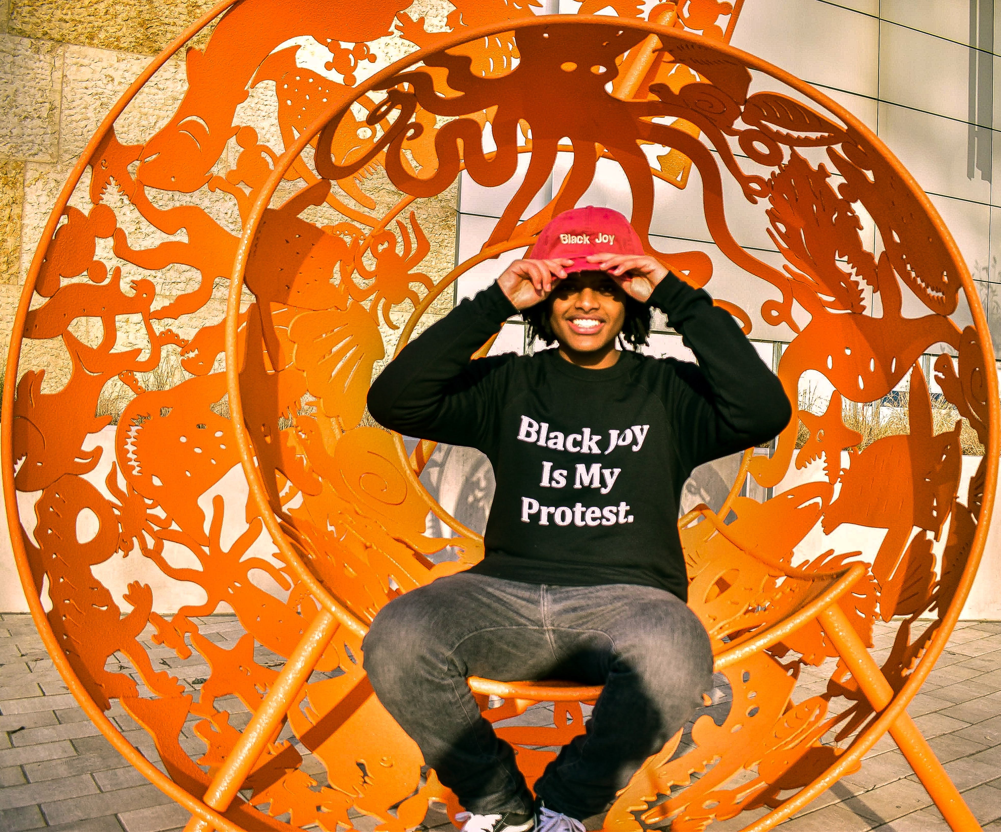 Young Black man wearing a "Black Joy Is My Protest" sweatshirt and a pink "Black Joy" hat.